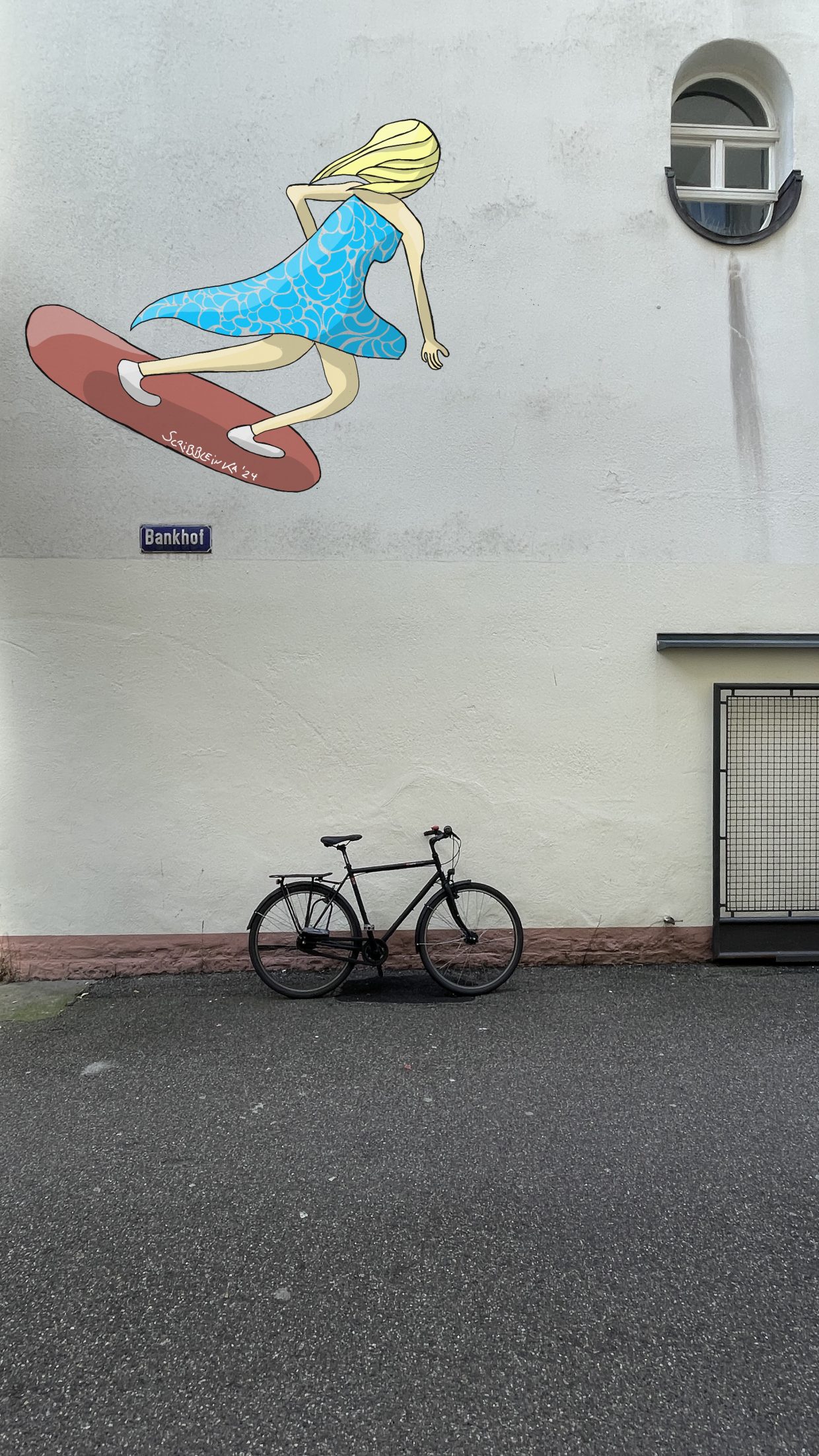Bankhof Karlsruhe. Eine Wand mit einem Fahrrad davor. An der Wand das Bild einer Frau in einem Kleid auf einem Board.