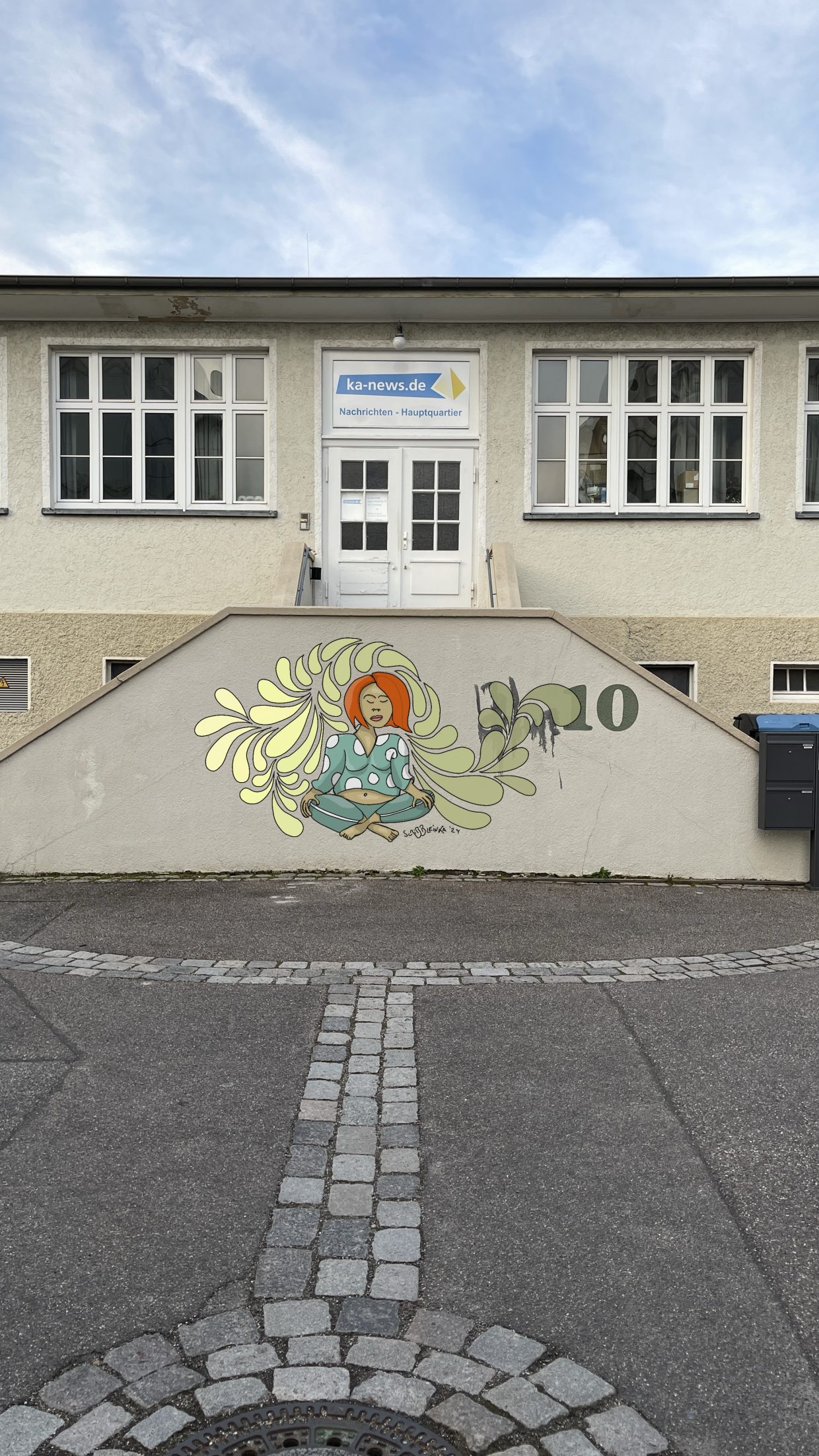 Treppe zum KA-News.de-Gebäude in der Karlsruher Oststadt. Davor ein Gemälde, auf der Mauer der Treppe mit einer meditierenden Frau in einer Art groben Gymnastik-Kleidung.