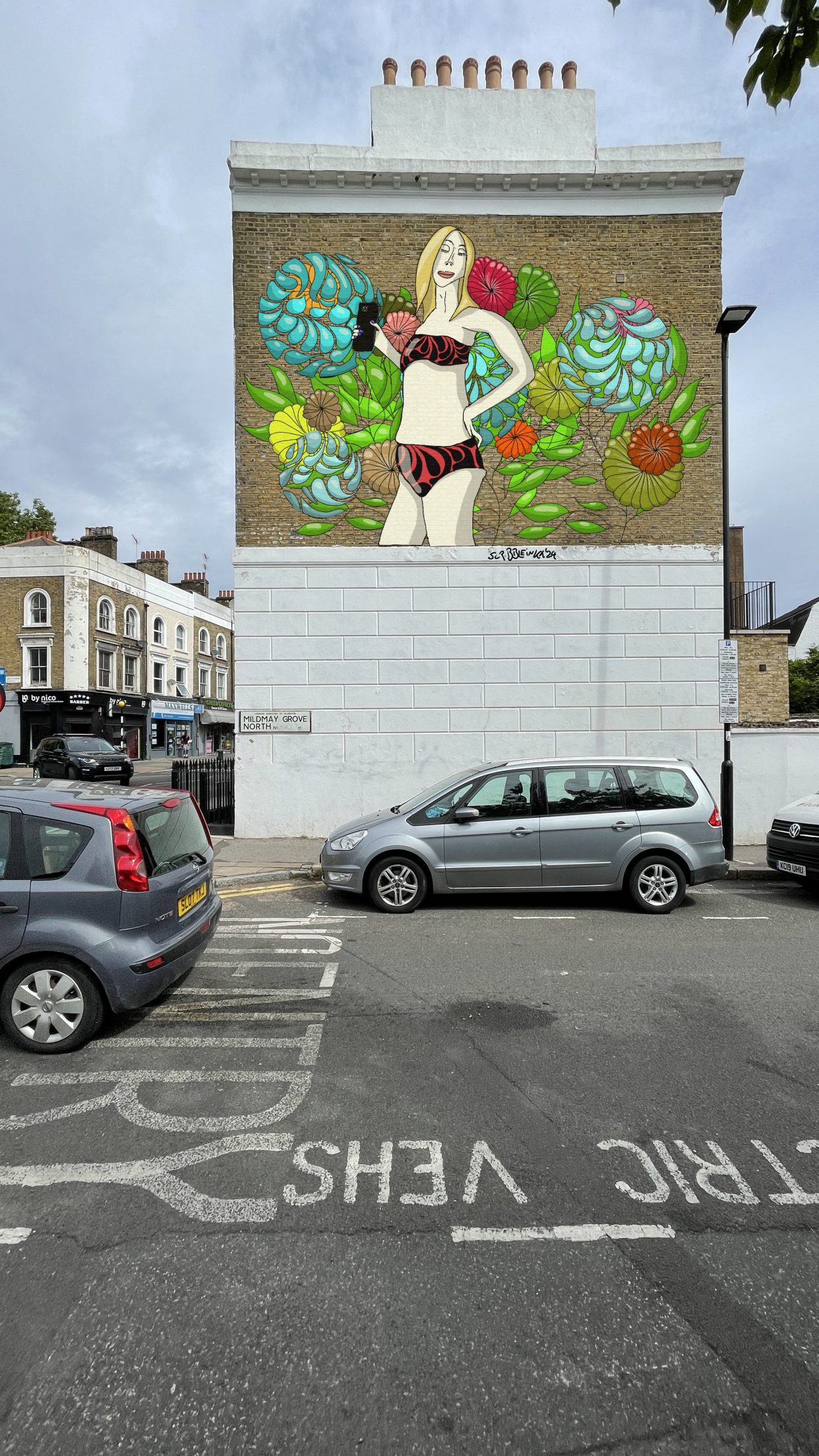 Parkplatz in London. Die Außenwand eines Hauses. Unten weiß. Oben ein Gemälde mit einer weißen Frau im Bikini und einem Handy in der Hand vor einer Blütenwand. Sehr vereinfacht, nicht fotorealistisch.